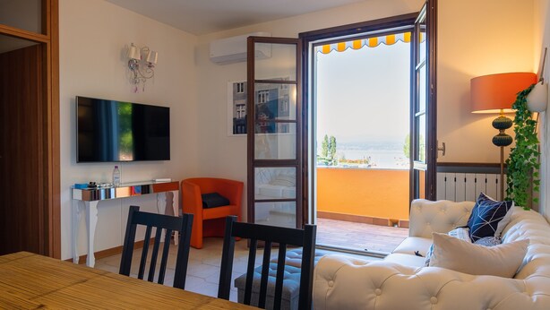 A living room with a TV, a dining table, chairs, a sofa, a balcony with a view, and a lamp. - La Casa sul Porto - Italian Homing (Sirmione)