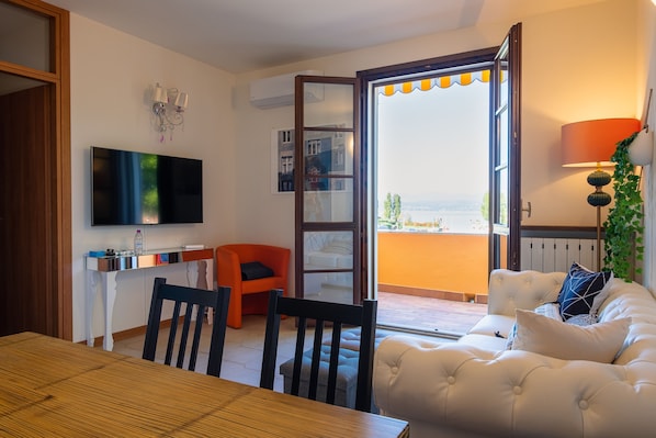 A living room with a TV, a dining table, chairs, a sofa, a balcony with a view, and a lamp.