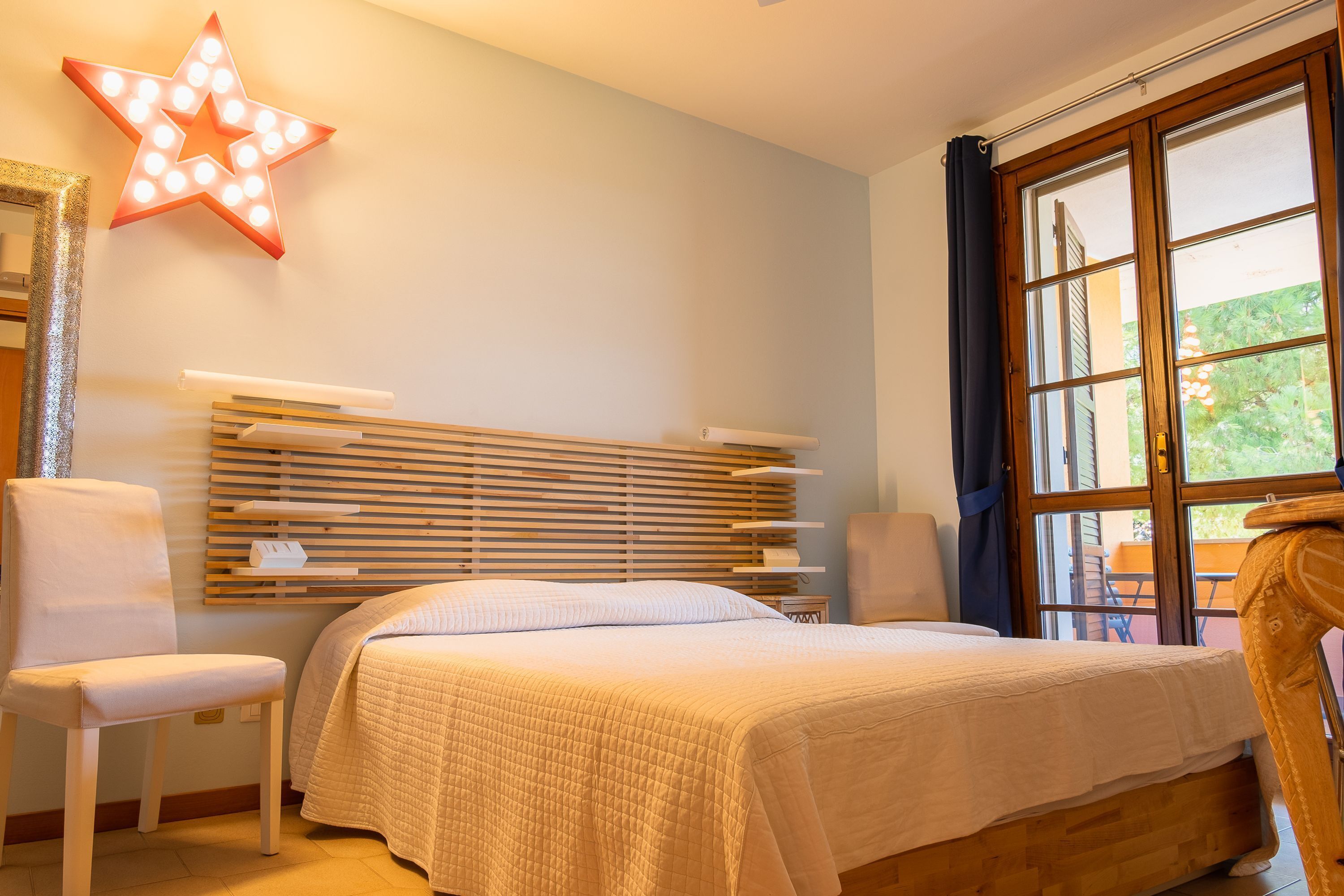 Une chambre à coucher comprenant un lit, une chaise, un luminaire en forme d’étoile, une fenêtre avec des rideaux et une tête de lit en bois.