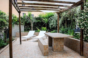 An outdoor patio with a stone barbecue area, two white lounge chairs, and a pergola structure.