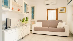 A modern living room with a white sofa, a TV on a white cabinet, a plant, and a window with a curtain. - La Casa di Nonna Agnese - Italian Homing (Padenghe sul Garda)