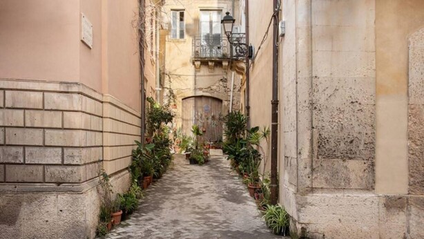 Une ruelle étroite, avec un chemin pavé bordé de bâtiments dotés de balcons et de plantes en pot.