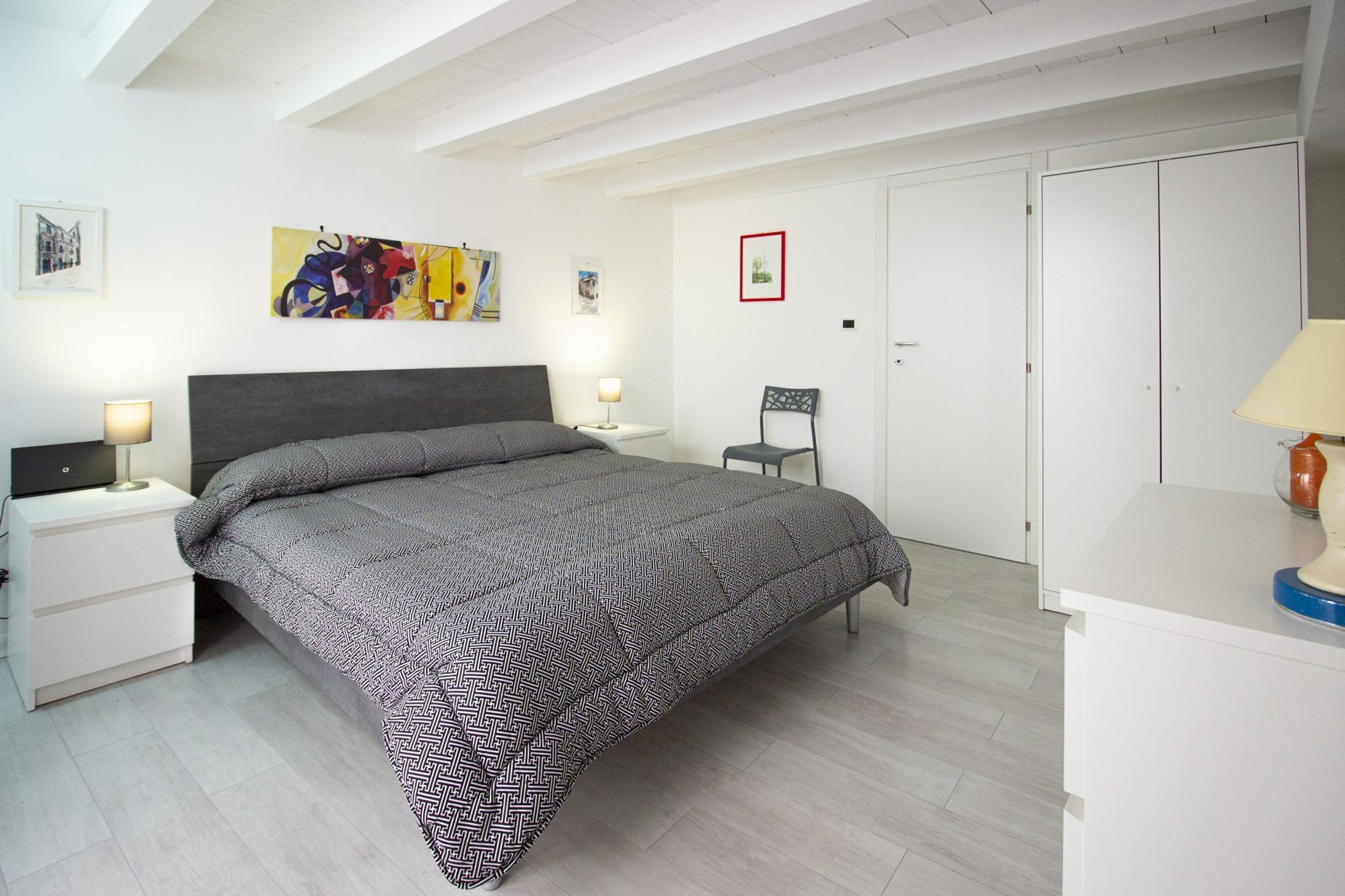 Un dormitorio con cama, mesitas de noche, una silla, armario y obras de arte en las paredes.