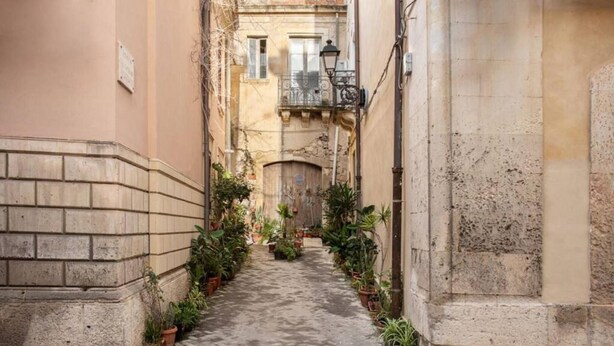 A narrow alleyway with potted plants lining the path, flanked by buildings with stone facades and a visible balcony.