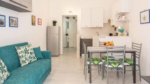 A compact living space with a teal sofa, a dining table with chairs, a kitchen area with white cabinets, and a refrigerator.