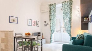 A dining area with a table and chairs, a bowl of fruit, and a window with patterned curtains.