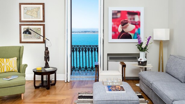 A living room with a view of the sea, a green armchair, a grey sofa, a small table with a lamp, and a framed map on the wall. - Aretusa Exclusive Sea View (Syracuse)