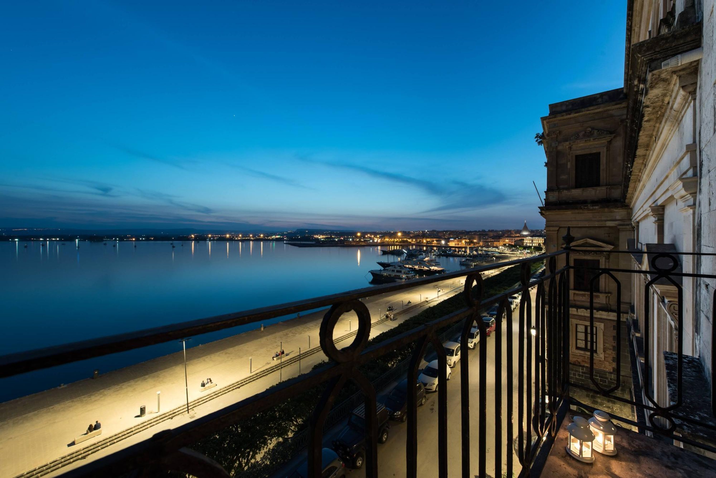 A waterfront view from a balcony with a railing, overlooking a calm river and a cityscape at dusk.
