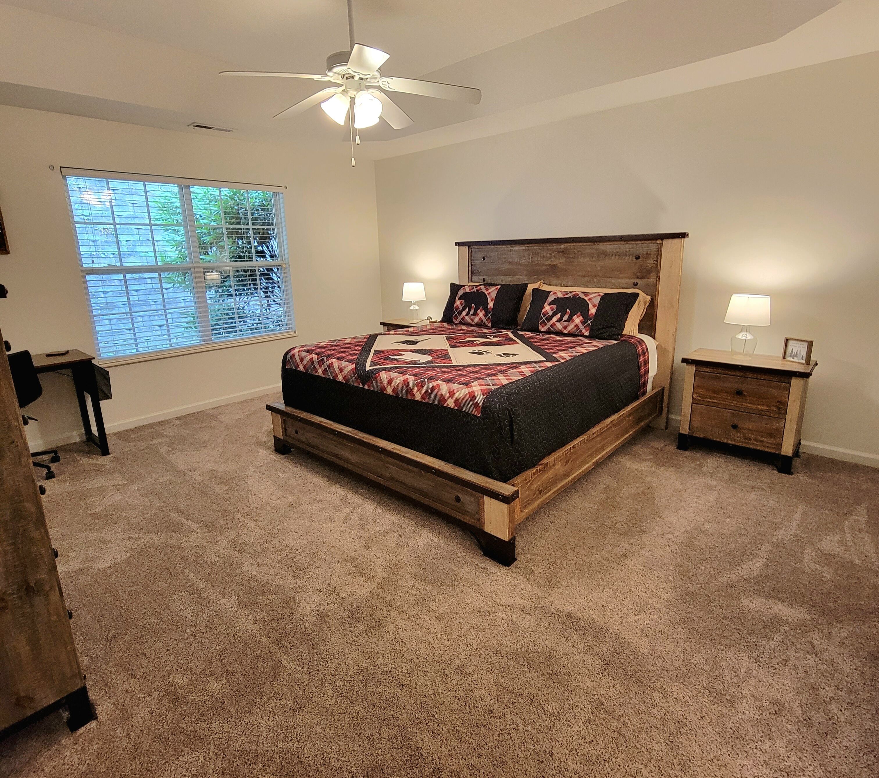 Une chambre avec un lit en bois, un ventilateur de plafond, une fenêtre avec des stores, un bureau et une table de chevet avec une lampe.