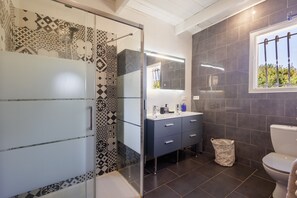 A modern bathroom with patterned tiles, a glass shower enclosure, a dark-toned sink area, and a window with a view.