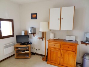 Quarto pequeno com televisão, abajur, armário e estante com cafeteira.