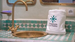 A bathroom with a gold faucet, a white towel with a logo, and a tiled backsplash. - Charming, quiet and comfortable Riad (Marrakech)