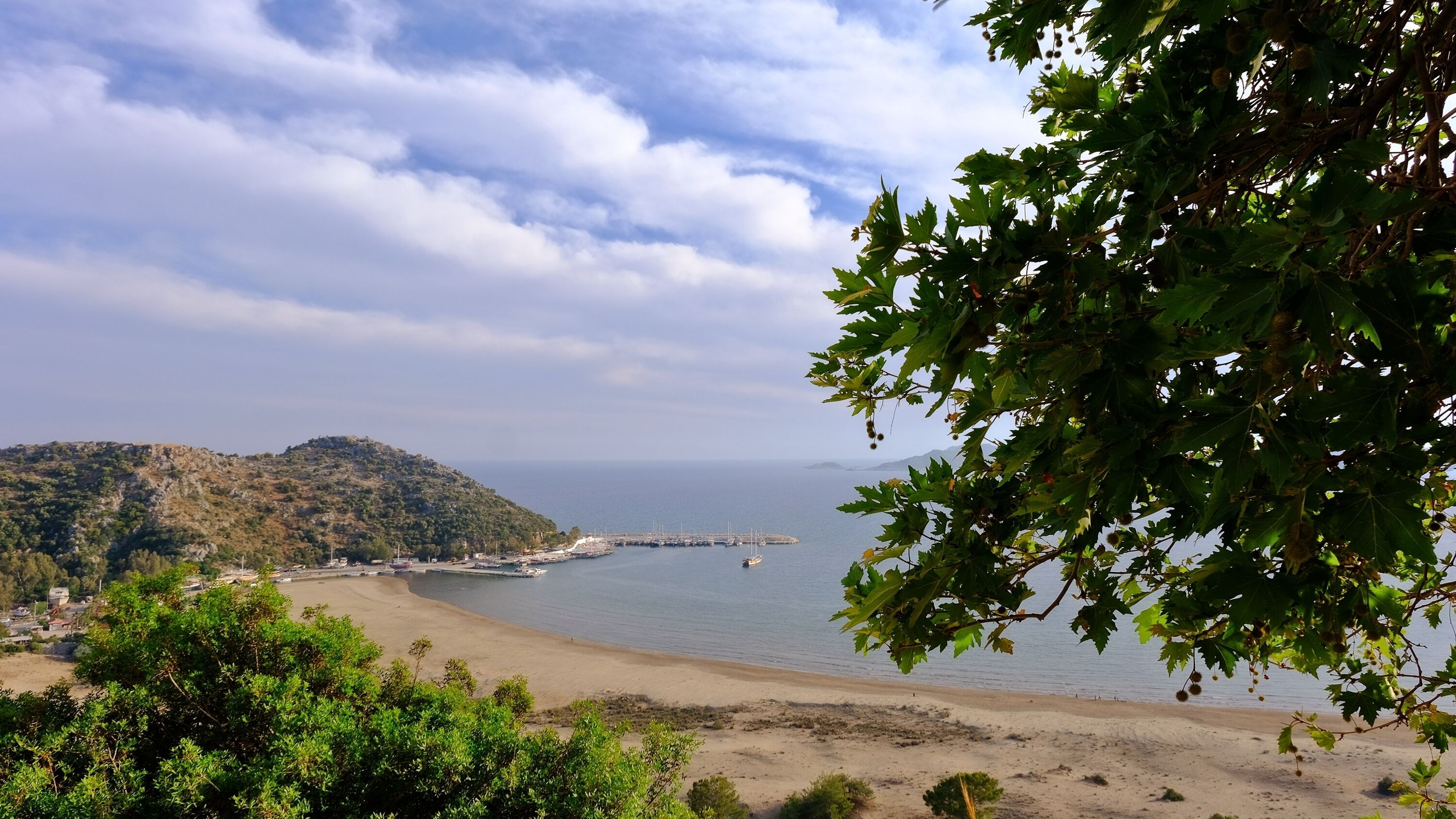 Un paisaje costero con una playa arenosa, un puerto deportivo y un fondo montañoso.