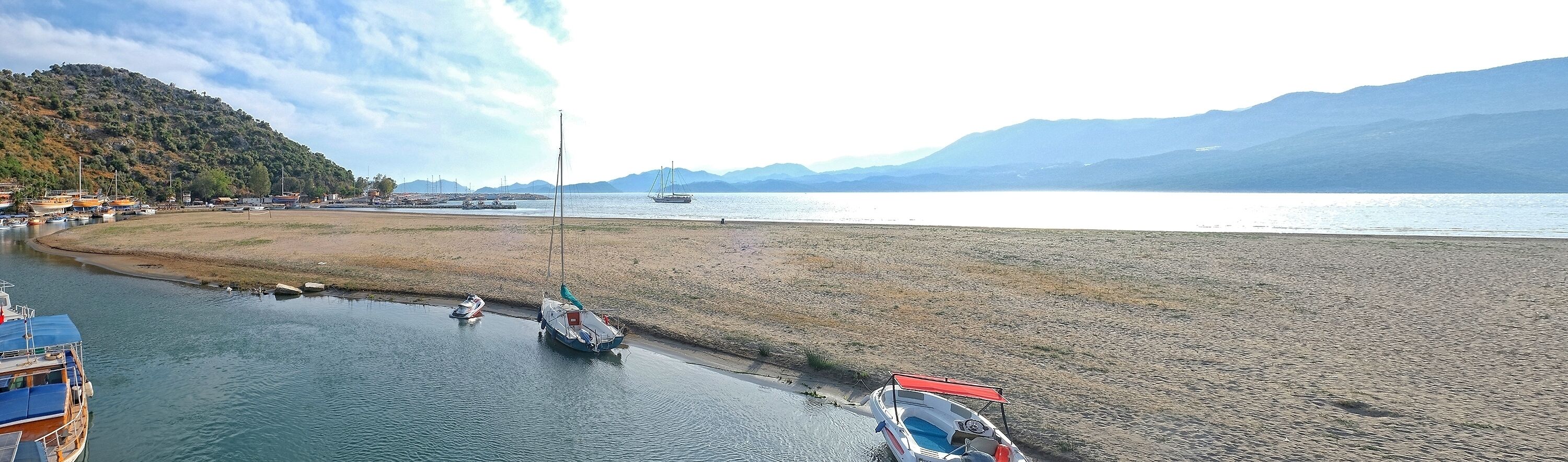 Una zona costera con barcos amarrados a la orilla, una playa arenosa y montañas al fondo.