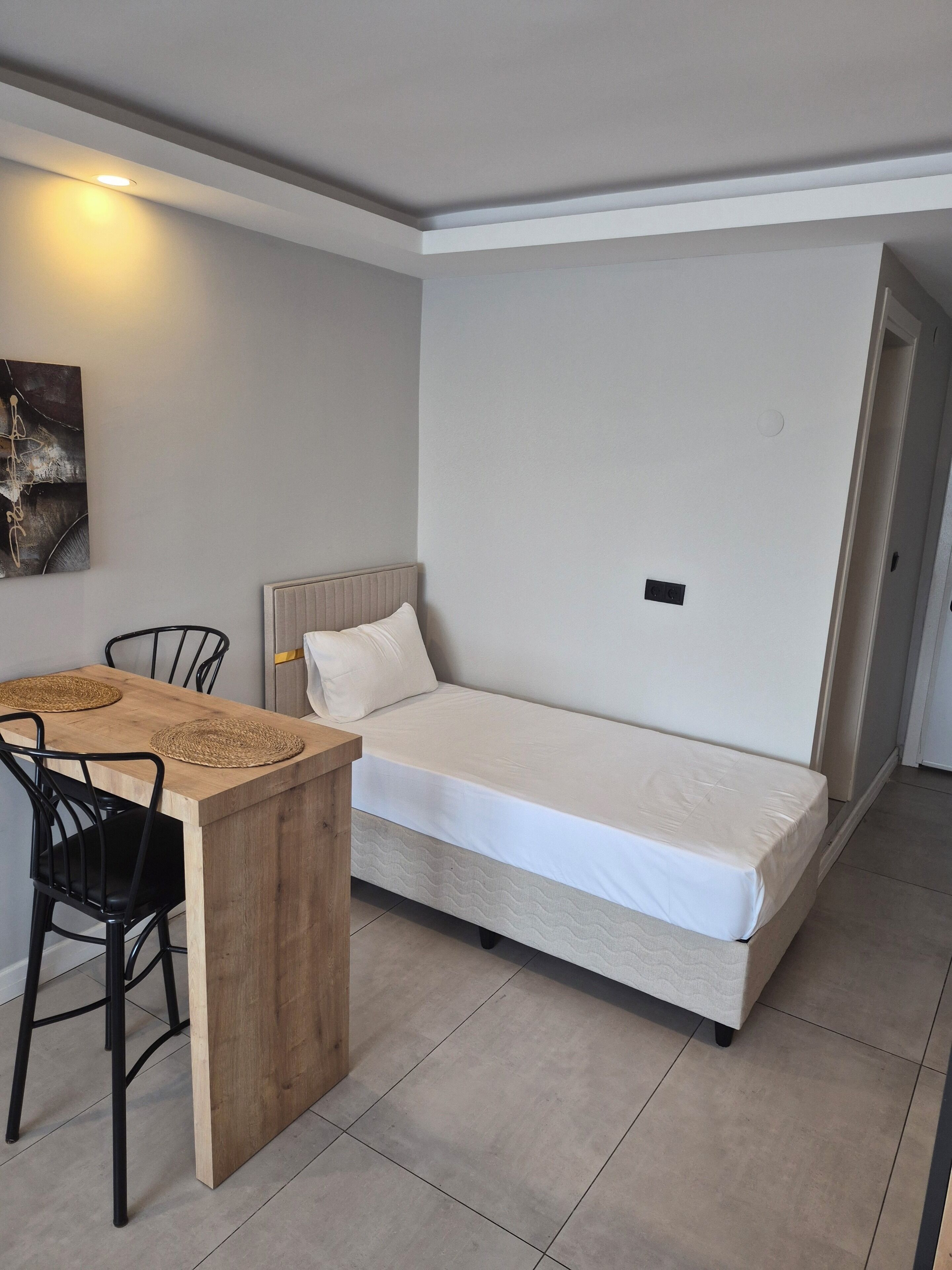 Una cama individual con cabecera, una mesa de madera con dos sillas, y una lámpara fijada a la pared.