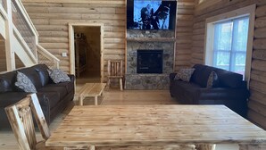Interior de una cabaña de troncos con chimenea de piedra, un televisor montado encima y dos sofás de cuero oscuro con cojines estampados, además de una mesa de centro de madera.