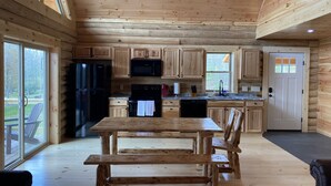 Interior de una cabaña de madera con cocina, mesa de comedor y ventanas amplias.
