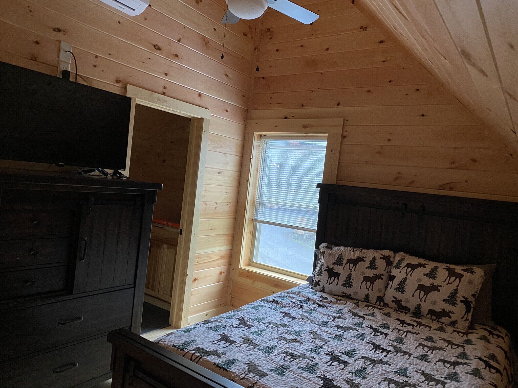 A bedroom with a wooden ceiling, a window with blinds, a bed with a moose-patterned pillow, and a dresser.