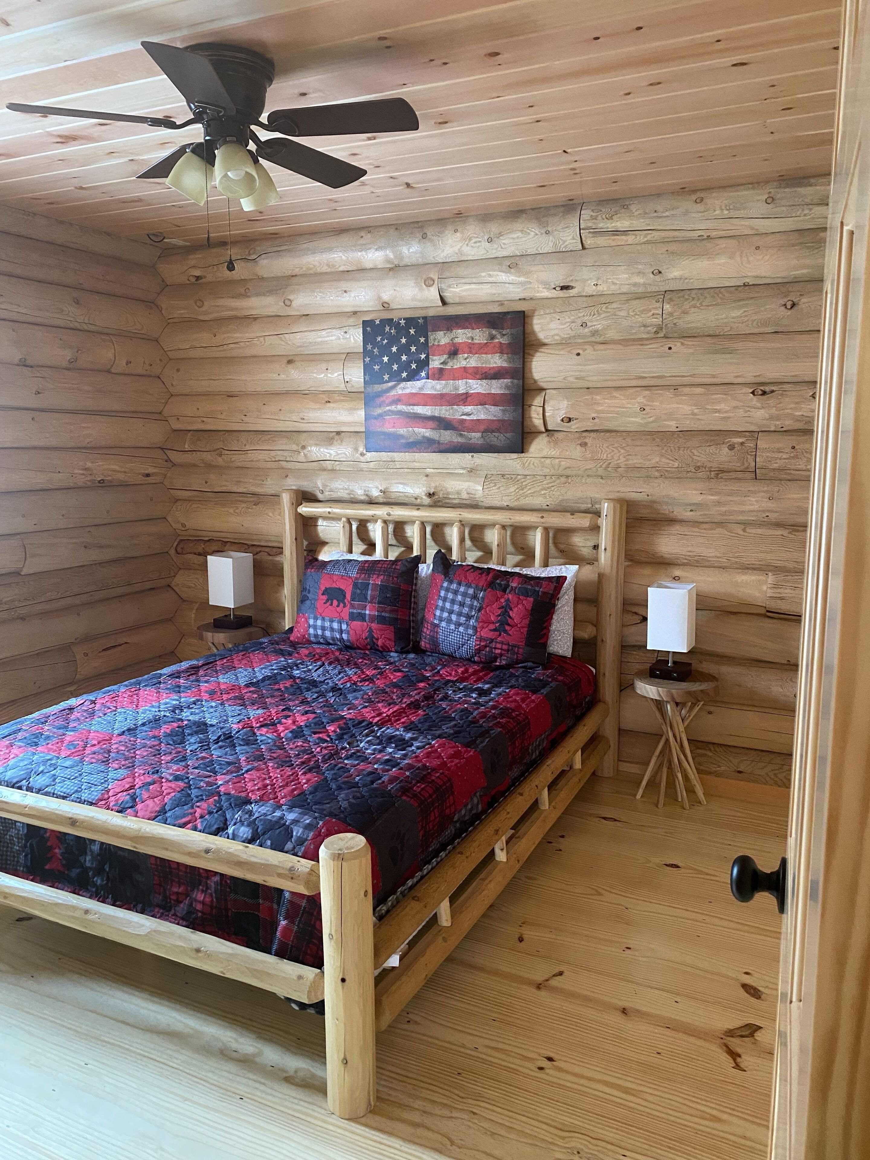 Une chambre à coucher de style cabane en bois, avec un lit, un ventilateur de plafond, un drapeau américain accroché au mur et une petite table de chevet avec une lampe.