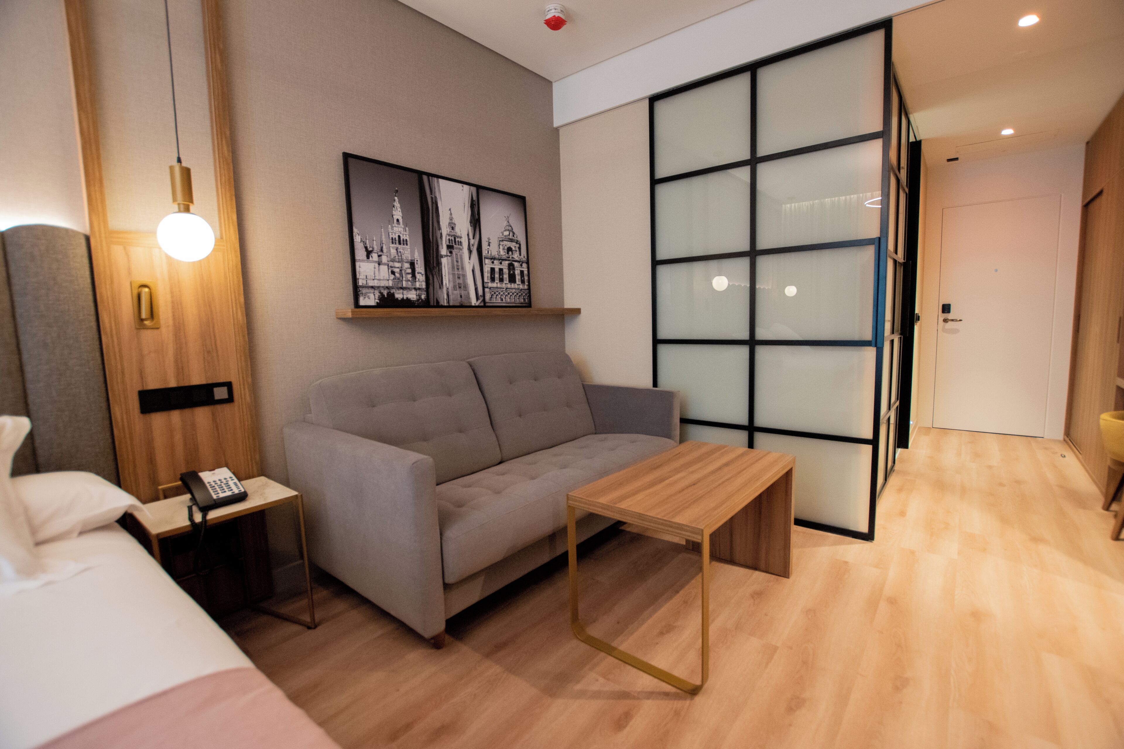 A modern hotel room with a sofa, a small wooden table, a bedside table with a phone, a wall-mounted light, and a framed cityscape picture.