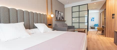 Una habitación de hotel moderna con una cama grande, un área de descanso con una silla y una mesita, y un ventanal amplio.