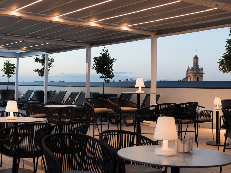 A rooftop terrace with outdoor seating, a view of a distant building, and a lamp on each table.