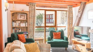 A cozy living room with a green sofa, a green armchair, and a wooden bookshelf filled with books. A television is on a wooden stand, and a window offers a view of the outside. - Byre Cottage, a Lake District cottage on a working farm (Broughton-in-Furness)