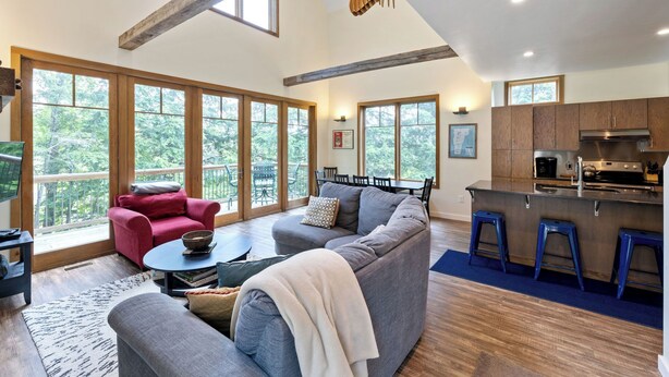 A spacious living area with a large gray sectional sofa, a red armchair, and a coffee table. There is a kitchen area with wooden cabinets and a stainless steel range hood. Large windows provide a view of the outdoors. - Mountainside 3-BR Ski-In/Out Okemo Chalet (Ludlow)