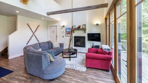 A spacious living room with a gray sectional sofa, a red armchair, and a fireplace. A staircase with wooden beams leads to an upper level. - Mountainside 3-BR Ski-In/Out Okemo Chalet (Ludlow)