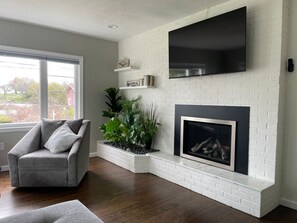 A living room with a fireplace, a grey armchair, and a large window. - Luxurious-Modern feel w/ custom features - AC, short walk to the Beach and parks (Waterford)