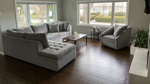 A living room with a grey sectional sofa, a small coffee table, and a grey armchair. There are two windows providing a view of a residential area.
