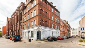 Un immeuble en briques de plusieurs étages, doté d’une façade blanche, avec des voitures garées le long de la rue et un panneau de signalisation bleu indiquant un parking.