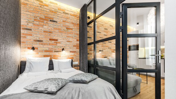 Une chambre moderne avec un mur en briques, un grand lit avec une literie blanche et une porte-fenêtre coulissante donnant sur un espace cuisine.