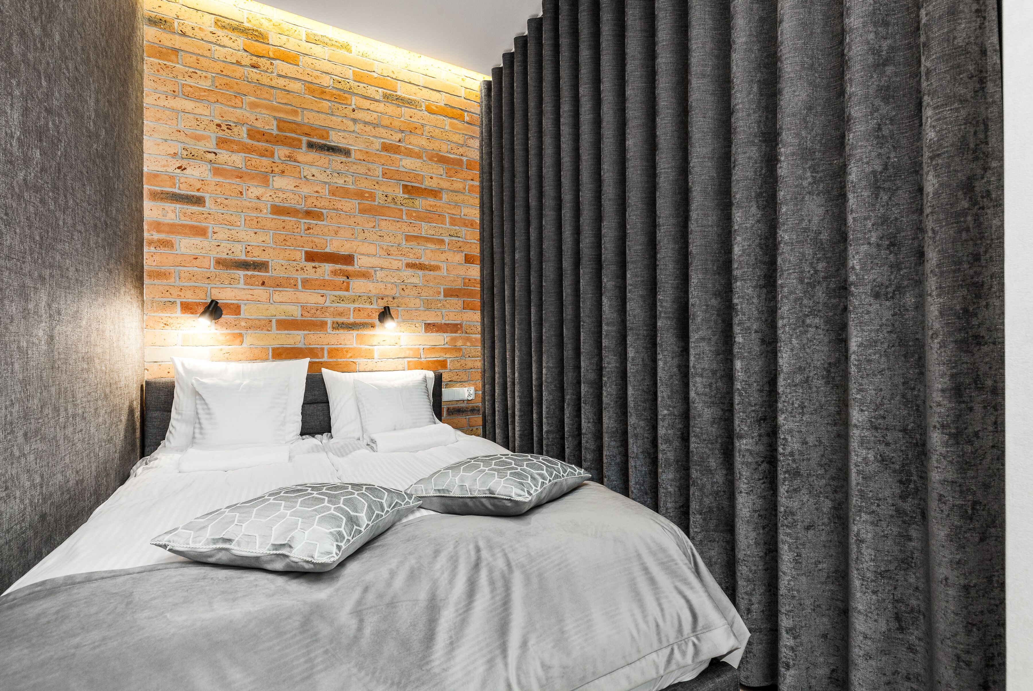 A modern bedroom with a brick wall, a bed with white bedding, and grey curtains.