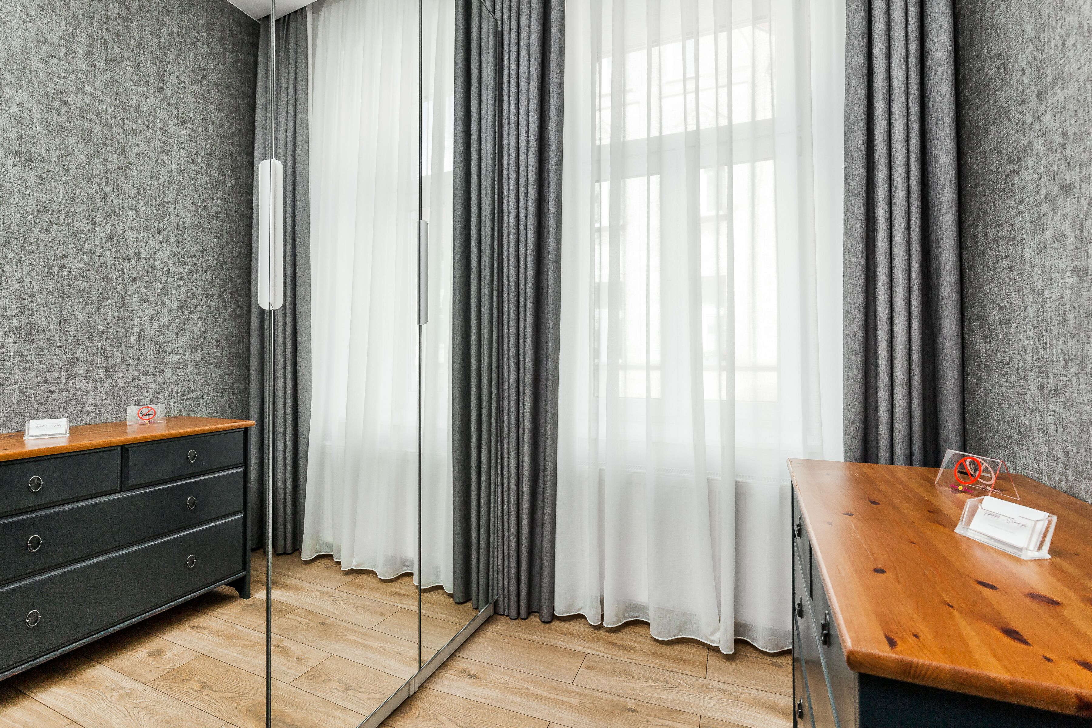 Une chambre d’hôtel avec une commode en bois, un miroir, une table en bois et une vue à travers des rideaux transparents.