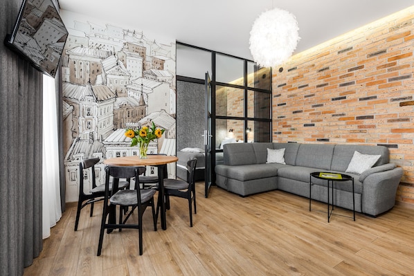 Uma sala de estar moderna com um sofá cinza, uma mesa redonda de madeira e cadeiras pretas. Uma janela grande com cortina branca é visível, e uma parede de tijolos se destaca.