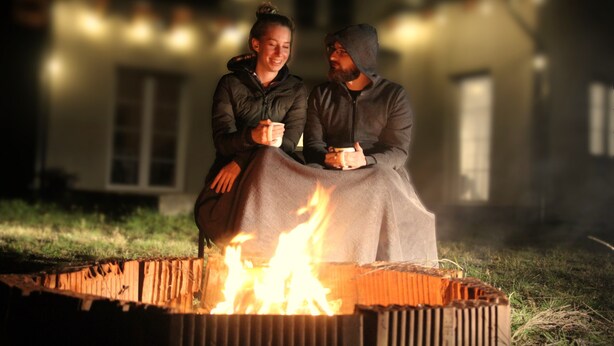 Two people sitting by a campfire in an outdoor setting with a house in the background.