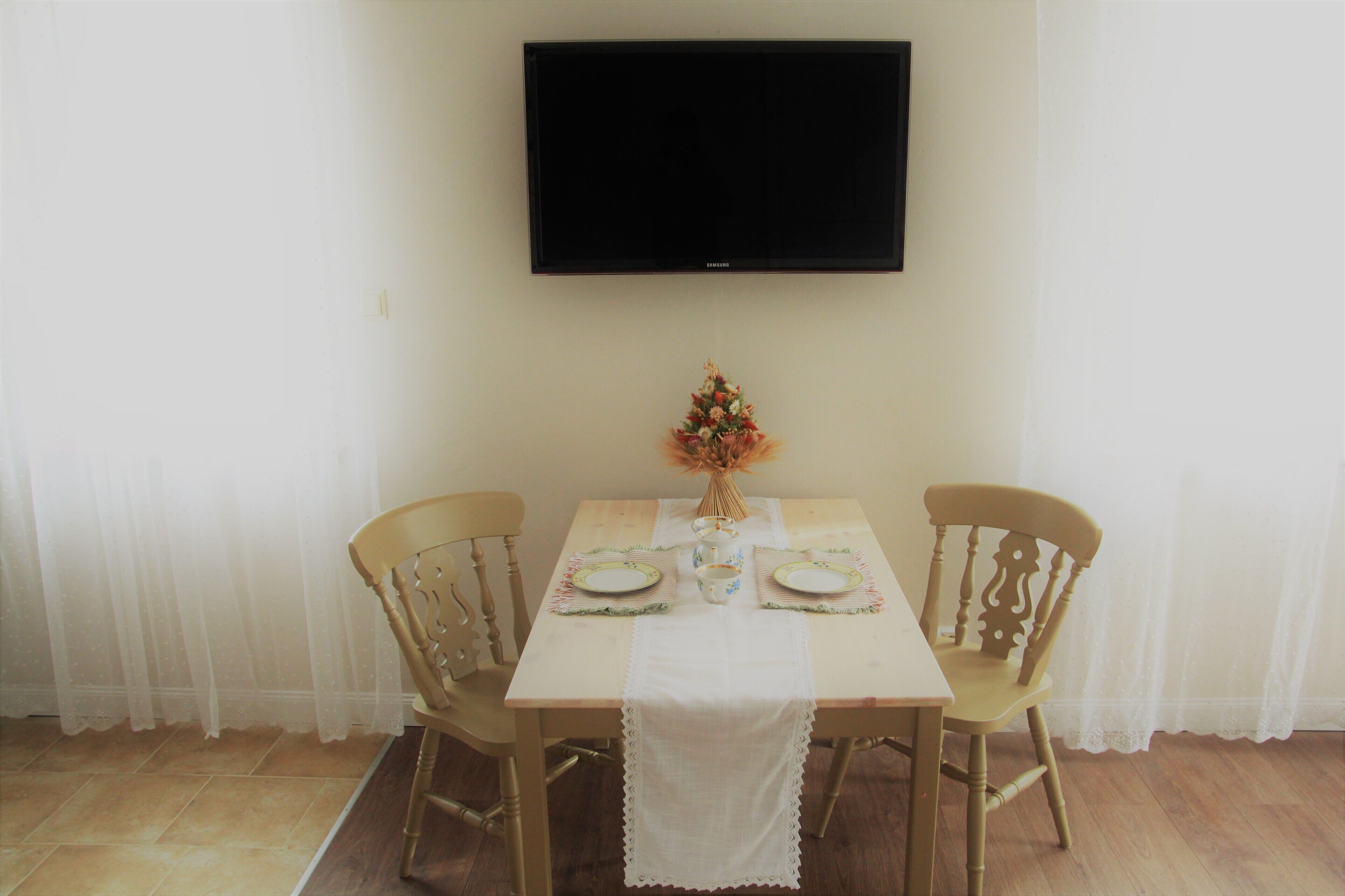 Uma área de refeições com uma mesa posta para dois, uma televisão montada acima e um vaso com flores na mesa.