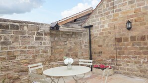 Un patio de paredes de piedra con una mesa redonda y sillas blancas, una pequeña construcción tipo casa y un farol.