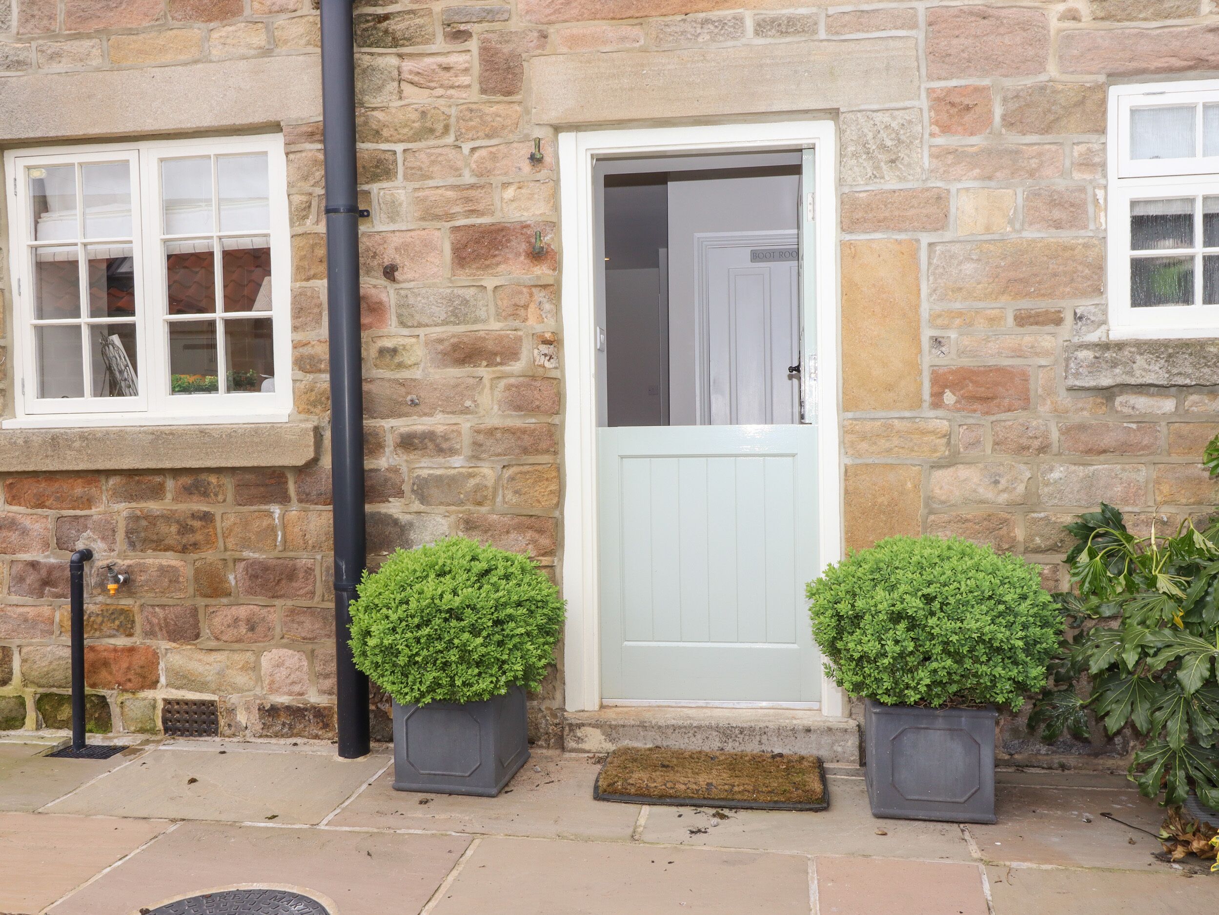 A stone building with a white door, two potted plants, and a metal grate on the ground.
