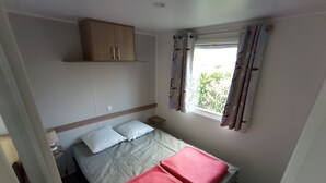 Ein kleines, ordentlich eingerichtetes Schlafzimmer mit Bett, Kissen, Nachttisch, Schrank und einem Fenster mit Vorhängen.