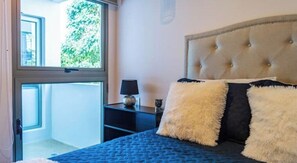 Ein Schlafzimmer mit einem gesteppten Kopfteil, einem Bett mit blauer Bettdecke und zwei flauschigen Kissen. Ein Nachttisch mit Lampe und einem kleinen Dekoartikel ist vorhanden. Ein Fenster mit Blick ins Grüne ist zu sehen.