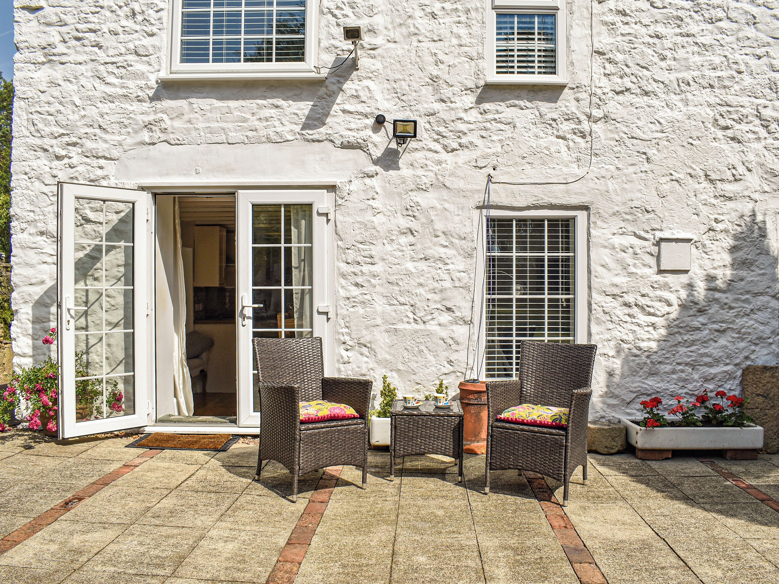 A white building with a stone facade, a glass door entrance, two wicker chairs with cushions, a small table, and potted plants.