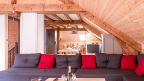 Une pièce chaleureuse, aménagée dans un style loft, avec un canapé gris foncé, des coussins rouges et un plafond en bois. On aperçoit à l’arrière-plan un coin repas avec une table et des chaises.