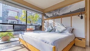 Une chambre moderne avec un grand lit, une fresque représentant un paysage urbain, une fenêtre donnant sur des immeubles et une plante en pot.
