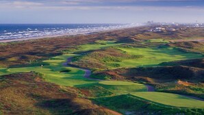 A golf course with a coastal view, a winding path, and a distant cityscape.