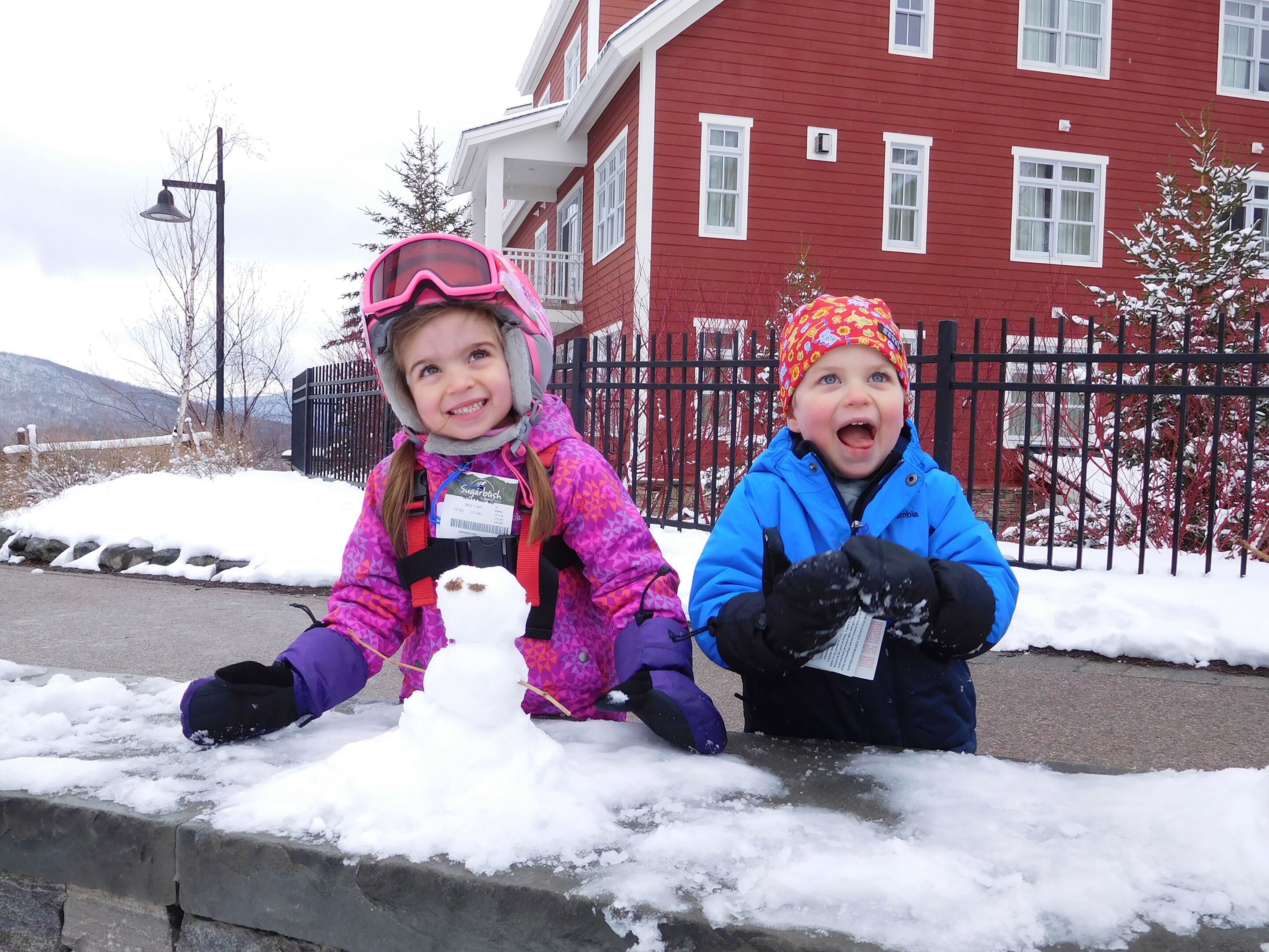 Dos niños construyen un muñeco de nieve frente a un edificio rojo con una valla negra.