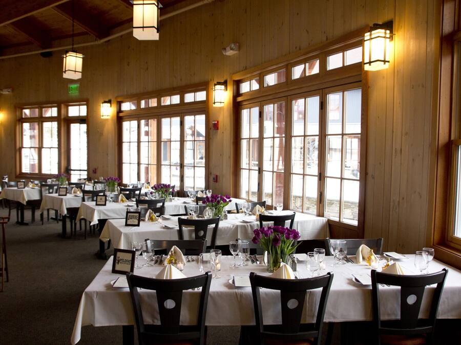 Restaurante com várias mesas preparadas para refeições, grandes janelas e luminárias pendentes.