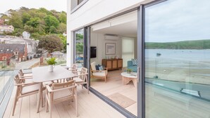 A modern hotel room with a large glass door leading to a balcony overlooking a beach. Inside, there is a dining table with chairs, a sofa, and a small side table with a plant.