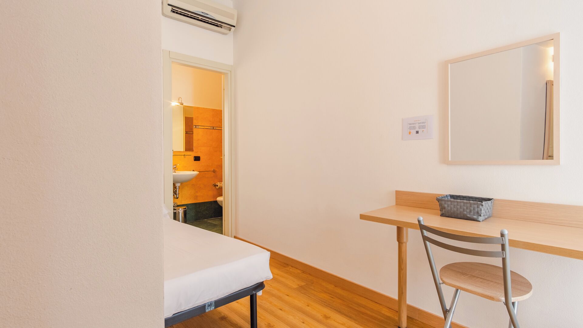 Een kleine, schone kamer met een bed, een bureau en een stoel. Er is een raam met uitzicht op een badkamer.